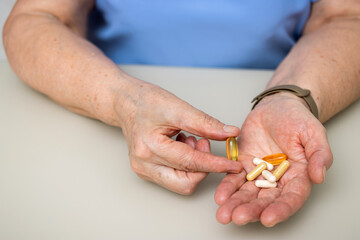 Senior woman with wrinkled old hands at the table holding Omega 3 capsule, vitamins and various pills for treatment. Healthcare and medicine concept