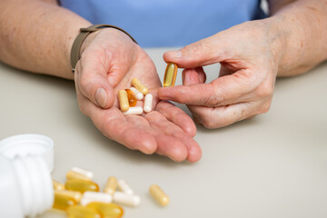 Senior woman with wrinkled old hands at the table holding Omega 3 capsule, vitamins and various pills for treatment. Healthcare and medicine concept