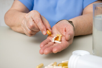 Senior woman with wrinkled old hands at the table holding Omega 3 capsule, vitamins and various pills for treatment. Healthcare and medicine concept