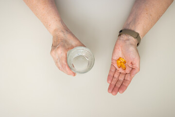 Senior woman with wrinkled old hands at the table holding omega 3 yellow capsules, fish oil pills and water glass. Healthcare and medicine concept, top view