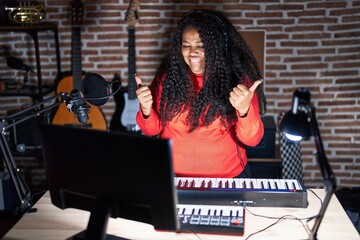 Plus size hispanic woman playing piano at music studio success sign doing positive gesture with hand, thumbs up smiling and happy. cheerful expression and winner gesture.