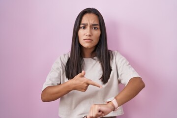Young hispanic woman standing over pink background in hurry pointing to watch time, impatience,...