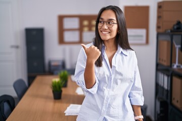Plakat Young hispanic woman at the office smiling with happy face looking and pointing to the side with thumb up.