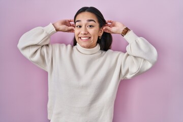 Young south asian woman standing over pink background smiling pulling ears with fingers, funny...
