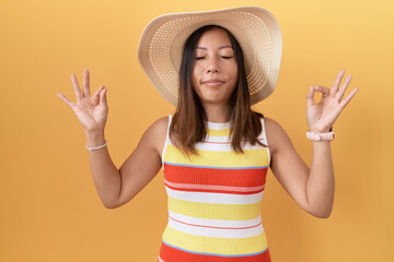 Middle age chinese woman wearing summer hat over yellow background relaxed and smiling with eyes closed doing meditation gesture with fingers. yoga concept.