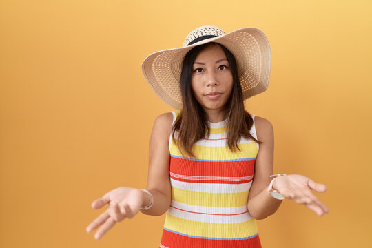 Middle age chinese woman wearing summer hat over yellow background clueless and confused with open arms, no idea concept.