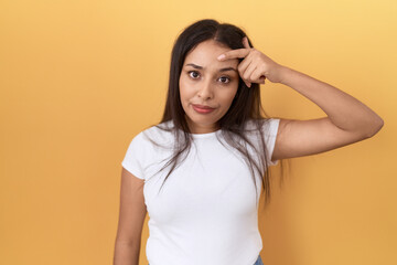 Young arab woman wearing casual white t shirt over yellow background pointing unhappy to pimple on...