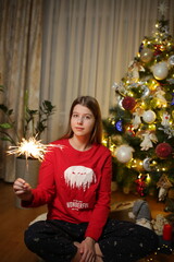 beautiful girl at home in pajamas sitting near the Christmas tree. red Christmas pajamas. the girl lit a sparkler near the Christmas tree. the magic of Christmas new year at home