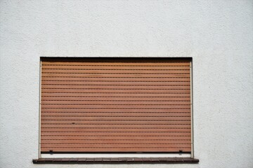 Weiße Hausfassade mit Fenster und runtergezogenen braun-roten alten Jalousien bei Sonne am Nachmittag im Frühling