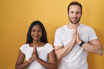 Interracial couple standing over yellow background praying with hands together asking for...