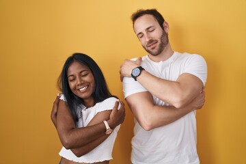 Interracial couple standing over yellow background hugging oneself happy and positive, smiling...