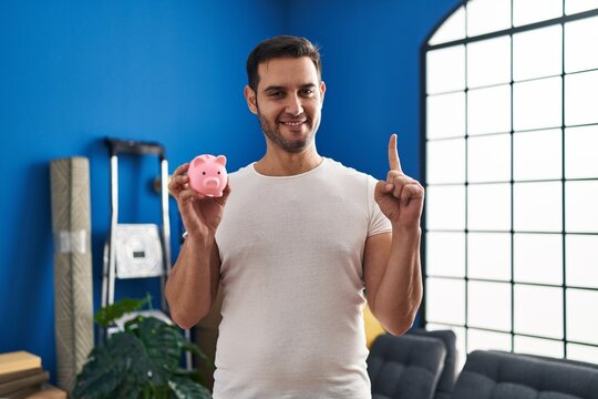 Young hispanic man with beard holding piggy bank at new home smiling with an idea or question pointing finger with happy face, number one