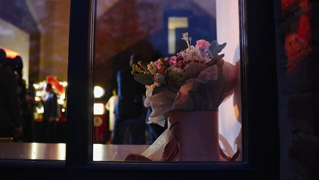 Bouquet of flowers in cardboard round box stands on windowsill outside window during corporate party. People in evening dresses are relaxing in decorated hall with multi colored stage lighting.