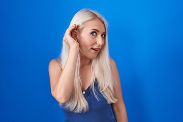 Caucasian woman standing over blue background smiling with hand over ear listening an hearing to rumor or gossip. deafness concept.