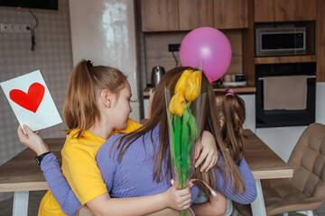 Daughters congratulate their mom on Mother's Day, a card with a heart, flowers and a balloon at home in the kitchen, they all hug. Children surprise their mother for the holiday.