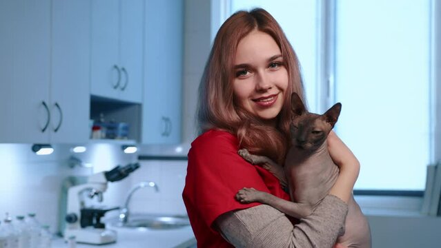 A young cute smiling female vet is petting adult Sphynx cat held in hands. The doctor is in a vet clinic in a modern office, wearing red doctor suit. High quality 4k footage