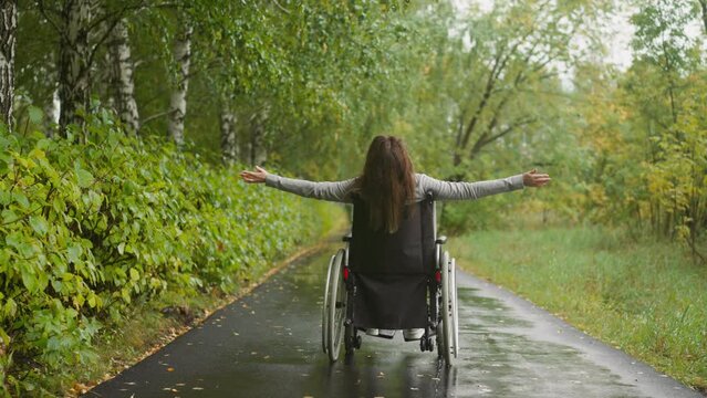 Woman with spinal cord injury stretches arms to sides enjoying walk in park on rainy day. Female recovers after accident resting in sanatorium backside view