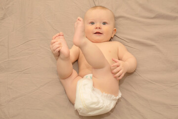 a baby with gray eyes in a diaper is lying in a crib, holding his legs with his hands and looking at the camera.