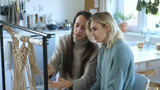 home courses, a woman teaches her friend how to weave macrame while sitting in the kitchen