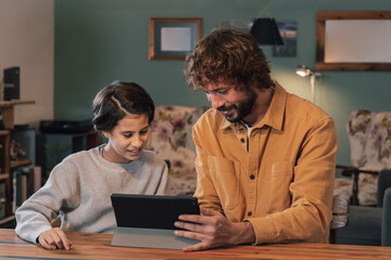 Young bearded man showing something on the tablet to a teenage girl at the dinner table at home
