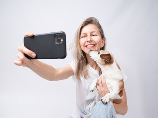selfie with blonde woman and her adorable small dog. Small dog Jack Russell terrier kissing blonde woman holding using cell phone. White clothes gray solid background. Social media content creator
