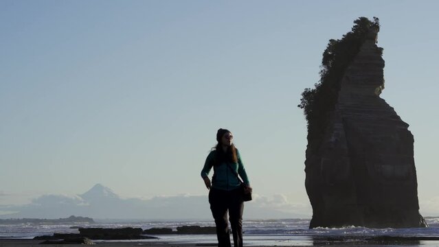 Brunette woman tourist walking by the beach of Three Sisters rock formation with Mount Taranaki behind. New Plymouth, New Zealand