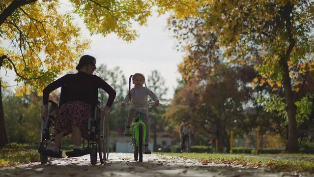 Aunt and niece spend time together. Young woman in wheelchair and preschooler girl riding bicycle rest in nature smiling contentedly. Active healthy recreation in park