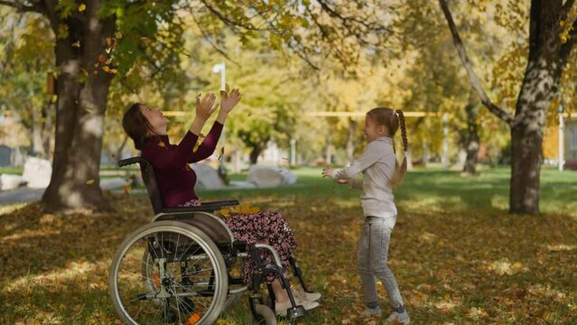 Girl with long braids throws up autumn leaves heap above mother in wheelchair. Couple raises hands laughing joyfully. Family recreation in nature