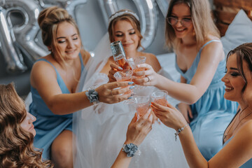 Young bridesmaids in blue silk dresses drink champagne in the bride's room. Beautiful women celebrating bachelorette party sitting on bed and with champagne.