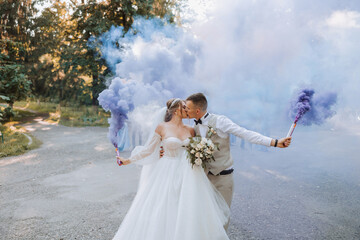 Fototapeta na wymiar In their hands, the newlyweds hold multi-colored smoke bombs of blue color. Groom and bride kiss. Wedding fun.