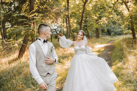A beautiful bride in a white dress with a long veil walks with a bouquet in the park with the groom. Young woman, art photo, wedding, bride, open shoulders. light.