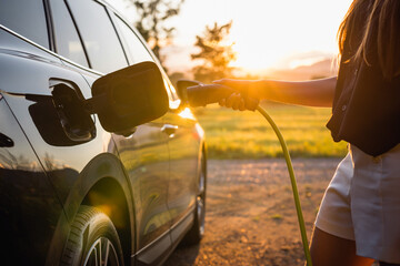 Female hand putting a cable charger in an electric car illuminated by the sun rays breaking through...