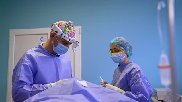 Surgeon in colorful cap works at thyroid operation. Doctor gives the used syringe to a nurse and takes new prepared one.