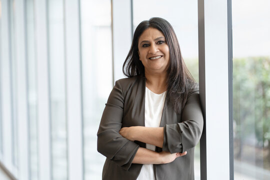 Confident smiling mature Indian businesswoman with arm crossed standing in modern office. Portrait of happy asian worker wearing stylish suit looking at camera indoors. Successful business concept 