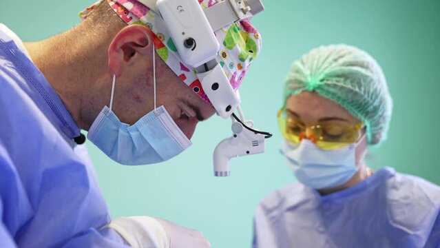 Side view of a surgeon in cap, mask and headlight who bent over the patient. Nurse in yellow protective goggles assisting the doctor.