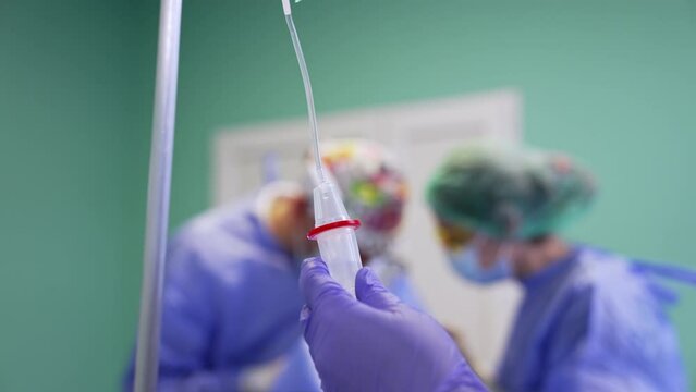Hand in blue latex gloves holding the reservoir of dropper. Arranging the drop counter during surgery. Surgeons work at backdrop in blur.
