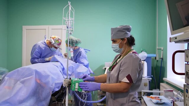 Nurse stands at the medical equipment arranging it for slow medicine delivery to the patient. Doctors at backdrop perform operation.