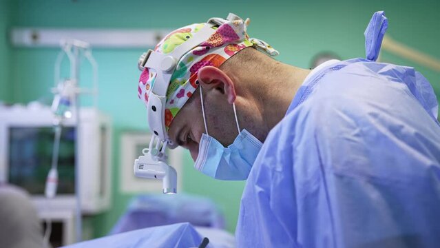 Surgery professional in mask, headlight and colorful cap. Side view. Portrait of a doctor focused on procedure.