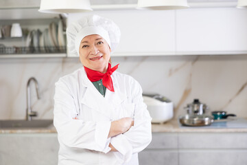 Elderly woman cook in chef hat and white cook uniform is standing at home in spacious kitchen and...