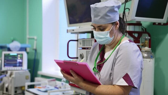 Nurse in surgery room wearing mask. Medic taking notes of the procedure carried out at the moment. Medical equipment at backdrop in blur.