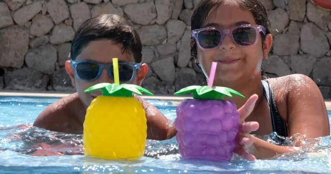 Cute 10 years old girl and 12 years boy swimming and racing for mocktails in colorful drinking cups and eye glasses in pool. Summer vacation concept: Front view, waist up wet child in 4k resolution