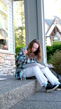 a girl draws with a pencil in a large notebook notebooks filming country house bottle of water white jeans black sneakers checkered blue jacket long brown hair of teenage girl