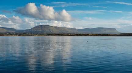 Panorama image on Benbulben mountains and blue water surface of Atlantic ocean with reflection of blue cloudy sky. Irish nature. County Sligo, Ireland. Popular tourist attraction. Stunning scenery.
