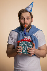 Happy young man wearing birthday hat cone blow pipe celebrating success win victory. Excited lucky guy in anniversary party isolated alone on beige studio background indoor. People lifestyles concept