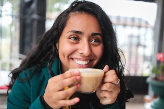 Headshot Of A Young Cheerful Hispanic Woman Smiling And Drinking Coffee In A Cafeteria