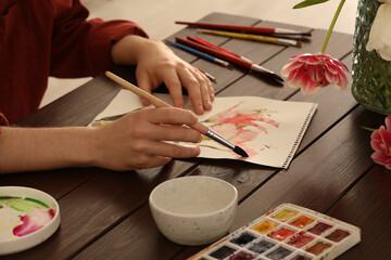 Fototapeta na wymiar Woman painting flowers with watercolor at wooden table, closeup. Creative artwork