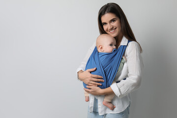 Mother holding her child in sling (baby carrier) on light grey background. Space for text