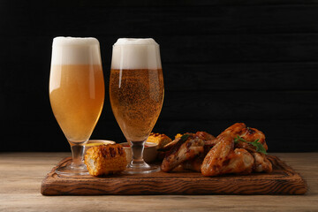 Delicious baked chicken wings, grilled corn and glasses with beer on wooden table against black background