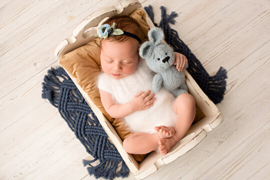 A cute newborn baby girl in a white jumpsuit with a flower bandage sleeps in a wooden crib on a brown pillow, top view. On a light wooden background. With a knitted blue bunny in the hands of newborn