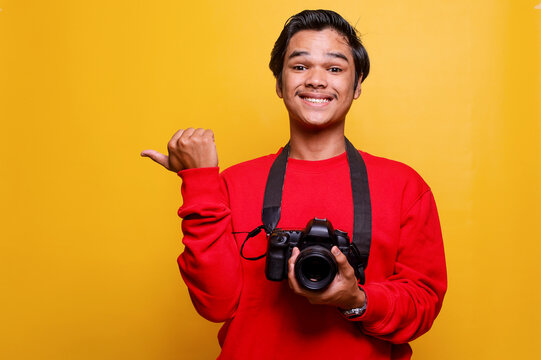Potrait of young professional photographer smiling and holds camera while pointing finger to the side isolated on yellow color background